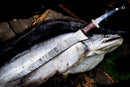 A freshly caught fish lies on a dark surface with a Texan Knives Handmade Damascus Fillet Knife 13” (Buffalo Horn, Rosewood & Brass Handle, Leather Sheath) blade gleaming atop it; an ornate green leaf sits nearby.