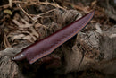 The Texan Knives Damascus Fillet Knife – 13" with a rosewood handle and leather sheath lies on a weathered tree trunk, surrounded by dried leaves and bark.