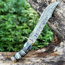 A 13" Vintage Damascus Steel Bowie Knife for Sale by Texan Knives, featuring a brass guard and camel bone handle, is displayed on a large tree branch with green foliage in the background.