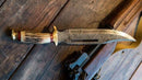 A Texan Knives Damascus Steel Vintage Bowie Knife, 13" with brass guard and stag horn handle, rests on a wooden surface next to a rifle; includes leather sheath.