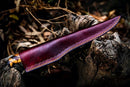 The Custom Handmade Damascus Steel Fillet Knife for Sale by Texan Knives, 13" long, features a patterned handle and rests on tree bark, partly sheathed in dark reddish-brown leather with yellow stitching, surrounded by dry leaves and twigs.
