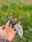 A hand holds the Texan Knives Premium Custom Handmade Damascus Steel Guthook Skinning Knife, featuring a 7.5” patterned blade and resin handle. Included is a leather sheath. The blurred background shows greenery.