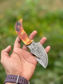 A hand holds the Texan Knives Custom Handmade Damascus Steel Guthook Skinning Knife, featuring a 7.5" patterned blade and camel bone handle, with blurred greenery in the background. Includes leather sheath.