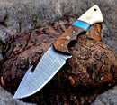 The Texan Knives Premium Damascus Guthook Skinning Knife features a patterned 10" Damascus steel blade and a rosewood & bone handle, displayed on rough wood with fur in the background.