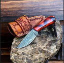 The Texan Knives Ranch Cowboy Knife features a Damascus steel blade and a red-brown resin handle, displayed on stone beside its engraved leather case, with rustic wood planks in the background.