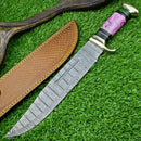 The Texan Knives Damascus Steel "The Bowie Knife" features a 16" patterned blade, brass guard, and a purple and gold resin pakkawood handle. It rests on green grass next to its brown tooled leather sheath.