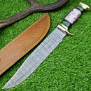 The Texan Knives Damascus Steel "The Bowie" Knife, 16" with brass guard and resin pakkawood handle, sits on artificial grass beside its brown leather sheath and an antler piece.