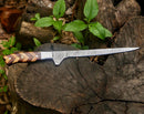 The Texan Knives Handmade Damascus Fillet Knife 8” with a rosewood and olive wood handle and leather sheath rests on a weathered tree stump outdoors among green plants.