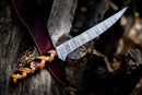 A Texan Knives Custom Handmade Damascus Steel Fillet Knife (13") with a multicolored wooden handle lies on a dark tree stump, its brown leather sheath partially visible in the background.
