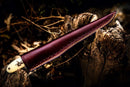 The Texan Knives Custom Handmade Filleting Knife for Fish features a 13" Damascus steel blade and a handle made of camel bone and rosewood, partially sheathed in dark brown leather, resting on a weathered tree stump among dry leaves.