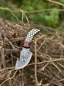 The Texan Knives Custom Handmade Damascus Steel Guthook Skinning Knife (7.5”) with a rosewood & resin handle and leather sheath is showcased on dry branches, with blurred green foliage in the background.