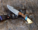 The Texan Knives Premium Damascus Guthook Skinning Knife features a 10” patterned blade and a rosewood & bone handle with blue inlay, resting on a leather sheath atop brown fur—a functional and striking hunting essential.