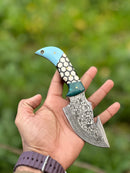 A hand holds the Texan Knives Custom Handmade Damascus Steel Guthook Skinning Knife (7.5”) with a blue and white resin handle, ornate blade, and leather sheath, set against a blurred green background.