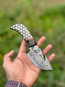 A hand holds the Texan Knives Custom Handmade Damascus Steel Guthook Skinning Knife with a patterned blade and a rosewood & resin handle, 7.5”, against a blurred green outdoor background. Leather sheath included.