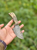 A hand holds the Texan Knives Custom Handmade Damascus Steel Guthook Skinning Knife with a sheep horn handle and patterned blade, 7.5”, shown outdoors with a blurred green background; includes leather sheath—great hunting gift.