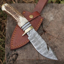 The Texan Knives Custom Handmade Damascus Steel Guthook Skinning Knife, 9" stag horn handle with brass guard and leather sheath, is displayed outdoors on a weathered wooden surface.