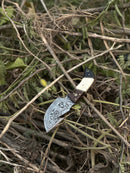 The Texan Knives Custom Handmade Damascus Steel Guthook Skinning Knife (7.5”) features a patterned blade and a rosewood, camel bone & black sheet handle, shown outdoors on dry branches and leaves. Includes leather sheath.