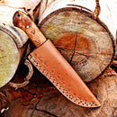 The Texan Knives Premium 10-Inch Damascus Steel Hunting Knife with Rosewood handle and a leather sheath is displayed on stacked logs, showcasing its rustic artisanal design—an ideal gift item.