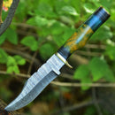 The Texan Knives Damascus Steel Hunting Knife – 10" features a patterned blade, a resin pakkawood handle with blue and yellow swirls, and a brass guard. Displayed outdoors among green leaves. Leather sheath included.