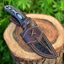 The Texan Knives Custom Handmade 9" Damascus Steel Hunting Knife with a black and gray G10 handle and leather sheath lies on a tree stump, framed by green foliage.