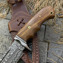 A close-up of the Texan Knives 🔥 Handmade Damascus Steel Hunting Knife with a polished rosewood handle and Damascus guard, showcased on its stitched leather sheath against gray stone tiles and dried plants.