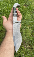 A hand holds the Texan Knives Custom Handmade 12" Damascus Steel Hunting Knife with a buffalo horn handle and Damascus steel bolster, shown outdoors; includes a leather sheath.