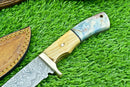 The Texan Knives Handmade Damascus Steel Hunting Knife, featuring an olive wood and resin handle, rests on green grass beside its brown leather sheath with yellow stitching.