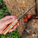 A hand holds the Texan Knives Custom Handmade 10" Damascus Steel Hunting Knife with an olive wood handle outdoors by a tree trunk, while its brown leather sheath rests on the tree.