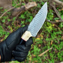 A gloved hand holds the Texan Knives Custom Handmade Skinning Knife, featuring a Damascus steel blade and a buffalo horn & camel bone handle. The background is an outdoor scene with green grass and scattered twigs.