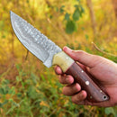 A hand holds the Texan Knives Custom Handmade Skinning Knife, featuring a Damascus steel blade and wood-bone handle, set against blurred green foliage outdoors.