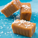 Three Saltopia Caramel Sugar: DATE NIGHT candies from SALTOPIA Infused Sea Salts rest on a blue surface, featuring a smooth, glossy texture and coarse sea salt topping. Visible ridges line the sides of these rich caramel treats.