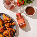 A plate of glazed chicken wings sits next to a jar of Honey Truck Co Raw Datil Pepper Infused Honey, a bowl of sauce, fresh parsley, and a jar of raw honey with a dipper, all arranged on a light-colored surface.