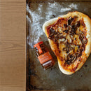 A baked pizza topped with mushrooms and cheese rests on a metal baking sheet beside a small jar of Honey Truck Co Raw Datil Pepper Infused Honey. Some flour is scattered across the baking sheet, and the left edge of the photo is framed by a wooden surface.