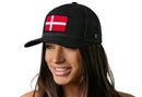 A woman with long brown hair smiles, wearing the Haka Hat Denmark Flag Trucker Hat—a black Danish snapback with a red and white embroidered Denmark flag patch—against a plain white background.
