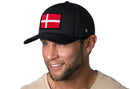 A man with a short beard smiles and looks to the side, wearing a Haka Hat Denmark Flag Trucker Hat—a black snapback by Haka Hat featuring an embroidered Danish flag patch on the front.