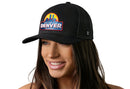 A woman with long brown hair smiles while wearing the Haka Hat Denver Mile High City Trucker Hat, a black cityscape snapback featuring a colorful 3D rainbow patch that says "Mile High City Denver Colorado." The background is white.