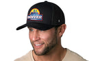 A man wearing the Haka Hat Denver Mile High City Trucker Hat, a black cityscape snapback with 3D embroidery and colorful skyline patch, smiles while looking slightly to the side.