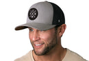 A smiling man wears the Haka Hat Denver Trucker Hat, a gray-black snapback with a DEN X circular logo, standing out against a plain white background.