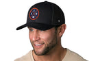 A man with a trimmed beard wears the Haka Hat Denver Trucker Hat | Black DEN X Football Snapback and a beige shirt. He smiles, looking slightly to the side, against a white background.