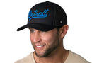 A smiling man in a beige shirt wears a Haka Hat Detroit Blue Script Trucker Hat (Black Michigan Snapback) against a plain white background.