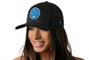 A smiling woman with long brown hair wears the Haka Hat Detroit Trucker Hat, a black DET X Football snapback by Haka Hat, featuring a blue circular logo with "DET" and a small car. The background is white.