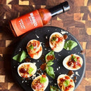 Deviled eggs topped with a tomato-bacon mixture, fresh herbs, and shredded cheese are arranged on a black plate. Nearby, a bottle of Bravado Spice Serrano & Basil Hot Sauce from Bravado Spice rests on a wooden surface, ready to bring botanical heat to every bite.