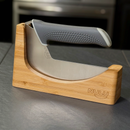 A curved-blade kitchen knife with a textured gray handle sits in the NULU Inc. Knife Holder Dock, crafted from acacia wood and labeled “NULU”, on a dark countertop—combining elegance with practical countertop knife storage.