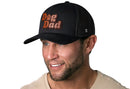 A smiling man with a trimmed beard and light shirt proudly wears the Haka Hat Dog Dad Trucker Hat Leather | Black Paw Snapback by Haka Hat, featuring bold orange and brown lettering and a paw print “o.”.