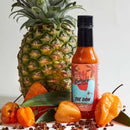A fresh pineapple, orange habanero peppers, green leaves, and Szechuan peppercorns are artfully arranged on a white surface alongside a bottle of Culture Hot Sauce The Don by Culture Hot Sauce (featuring the original recipe Pineapple Habanero Hot Sauce), all set against a plain white background.