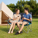 Two people relax outdoors on the DirectDeals Double Camping Chair with Carry Bag Cup Holders, smiling and enjoying red wine. A tent, green trees, and bottles of wine sit nearby.