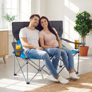 A smiling couple relaxes indoors on the DirectDeals Double Camping Chair with Carry Bag Cup Holders, drinks in hand, sunlight streaming in and a green potted plant nearby.