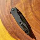 The Spaceman Knives Naiad EDC Pocket Knife by Spaceman Knives, closed and featuring a textured black handle, rests on a wooden surface with its titanium-plated blade concealed in the sunlight.