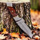 The Texan Knives Small Bushcraft Knife features a 9" Damascus steel blade, rosewood handle, and leather sheath, resting against a tree stump with colorful autumn leaves scattered around.