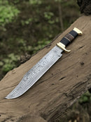 The Texan Knives Authentic Crocodile Dundee Knife features a 16” handmade Damascus steel blade, brass guard, and your choice of buffalo horn or leather handle with sheath, displayed outdoors on a weathered tree trunk.